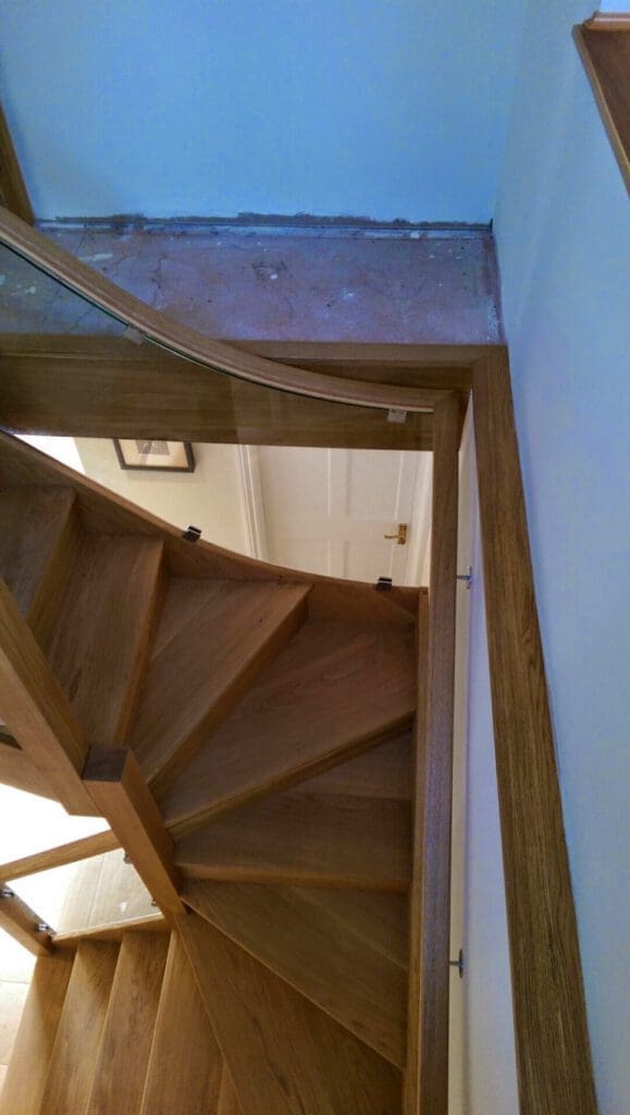 View from the top of a wooden spiral staircase with glass panel railings, leading down to a hallway with a closed white door; the top floor appears unfinished with exposed subfloor.