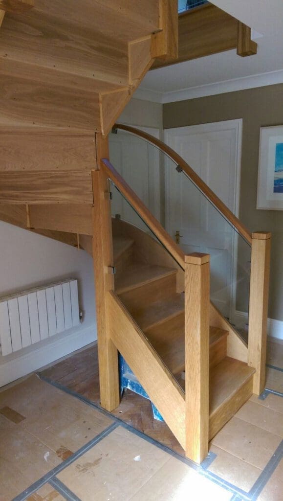 A modern wooden staircase with glass panel railings, set in a room with light-colored walls, a radiator, and a framed picture on the wall. The stairs have a sharp turn and unfinished floor protection.