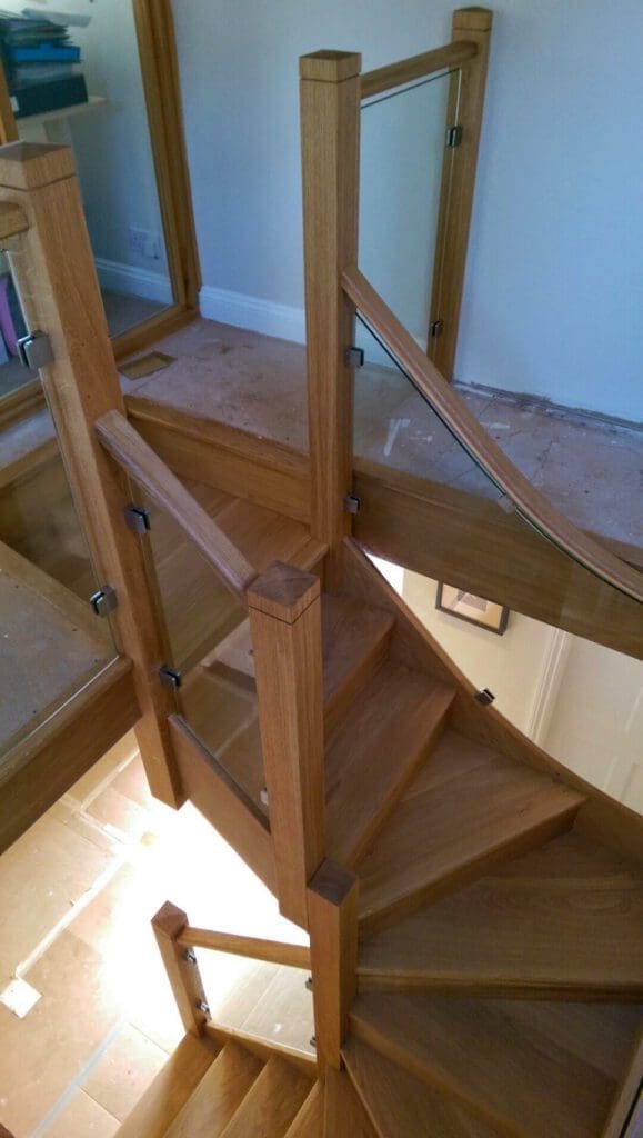 A modern wooden staircase with glass panel railings, featuring a turn and multiple landings. The stairs lead up from a tiled floor to a carpeted area. The design is angular with clean, straight lines.