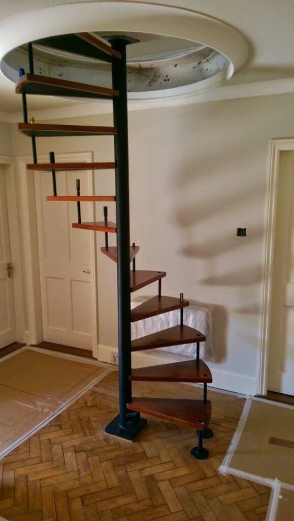 A modern spiral staircase with wooden steps and a black central pole leads up through a circular opening in the ceiling, set in a room with wooden parquet flooring and white walls.