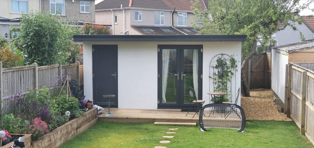 A modern garden shed with dark doors and windows sits at the end of a green backyard. There’s a small deck with a table and chairs, potted plants, a mini soccer goal, and flower beds along the fence.