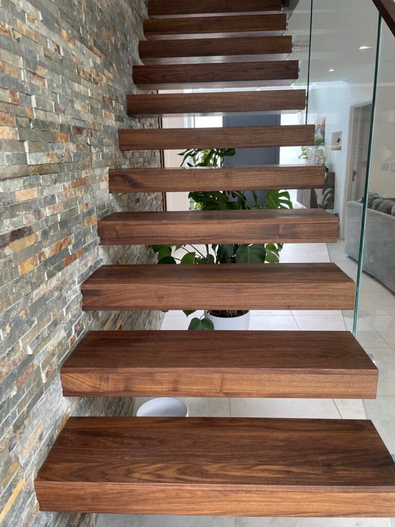 Floating wooden stairs with thick, dark planks mounted to a stone accent wall, leading upward beside a glass railing. A large green plant is visible at the bottom near a modern living area.
