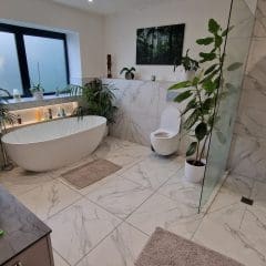 Modern bathroom with white marble tiles, a freestanding bathtub, glass shower, and toilet. Green plants and a large window add a natural touch. Soft bath mats are placed on the floor. A sink and countertop are visible.