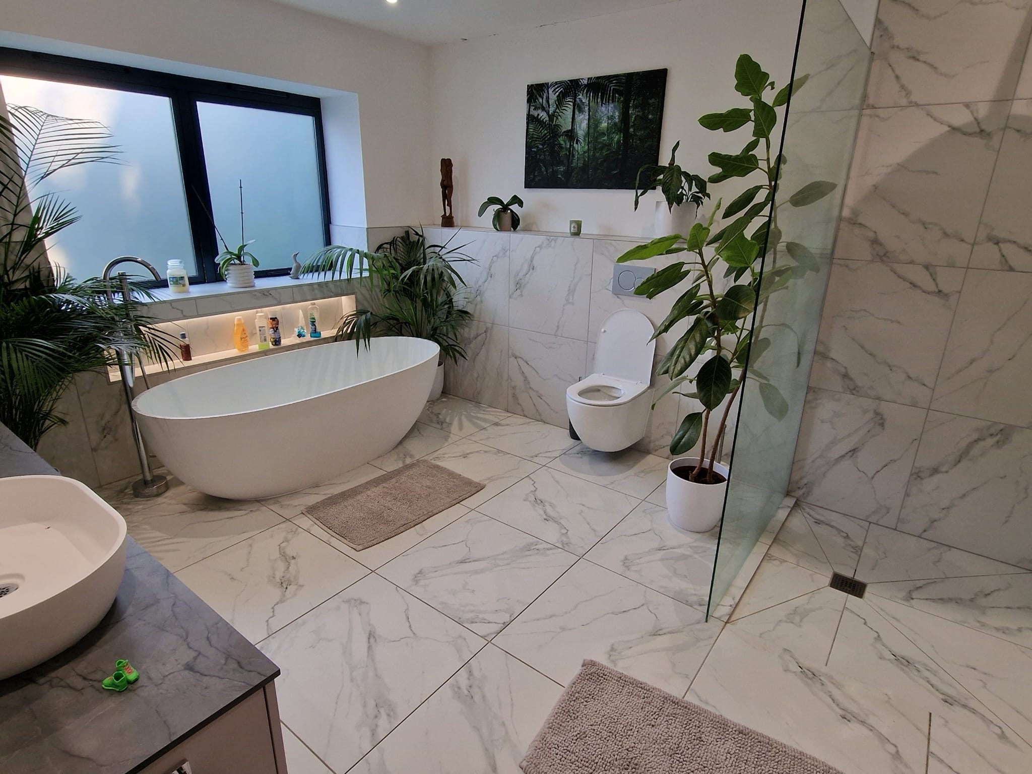 Modern bathroom with white marble tiles, a freestanding bathtub, glass shower, and toilet. Green plants and a large window add a natural touch. Soft bath mats are placed on the floor. A sink and countertop are visible.