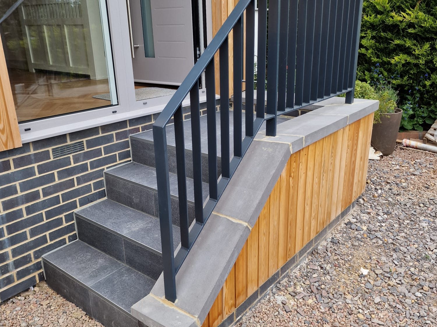 A small outdoor staircase with dark railings and gray tiles leads to a door on a modern house with wood and brick siding, set above a gravel ground.