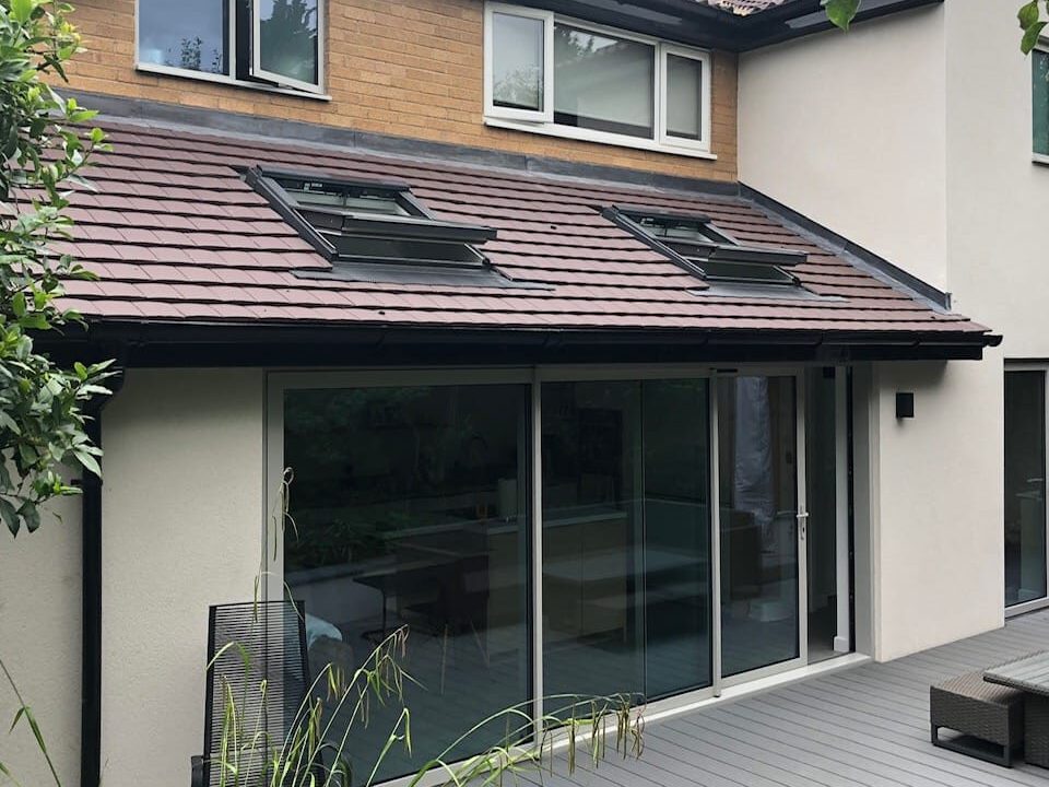 A modern two-story house with cream walls, brick accents, and brown roof tiles features large sliding glass doors and skylights, opening onto a gray deck with outdoor seating and surrounded by greenery.
