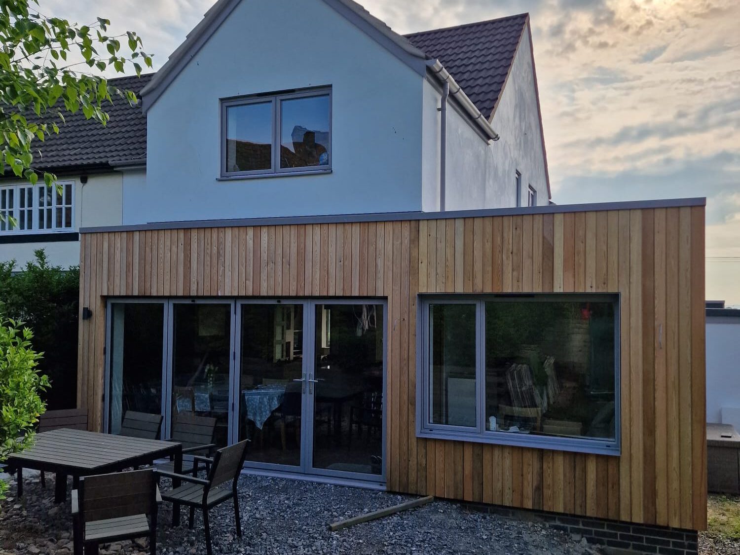 A modern house extension with vertical wooden cladding, large glass sliding doors, and a gravel patio area with outdoor furniture. The sun is partially visible through a cloudy sky above the house.