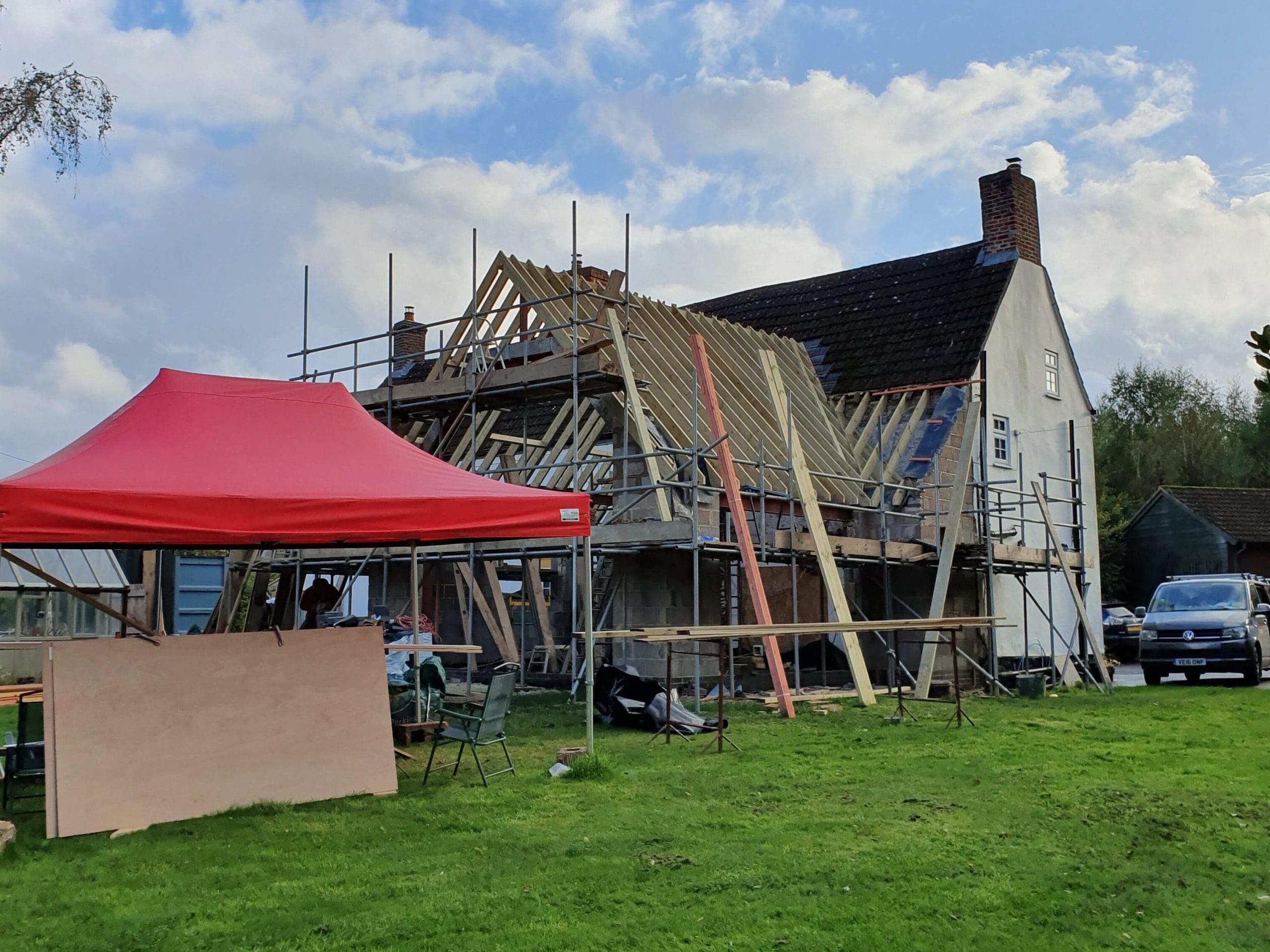 A house under construction with exposed roof beams and scaffolding, a red canopy tent, wooden boards, and tools on the grassy lawn. Two parked cars and a greenhouse are visible in the background under a partly cloudy sky.