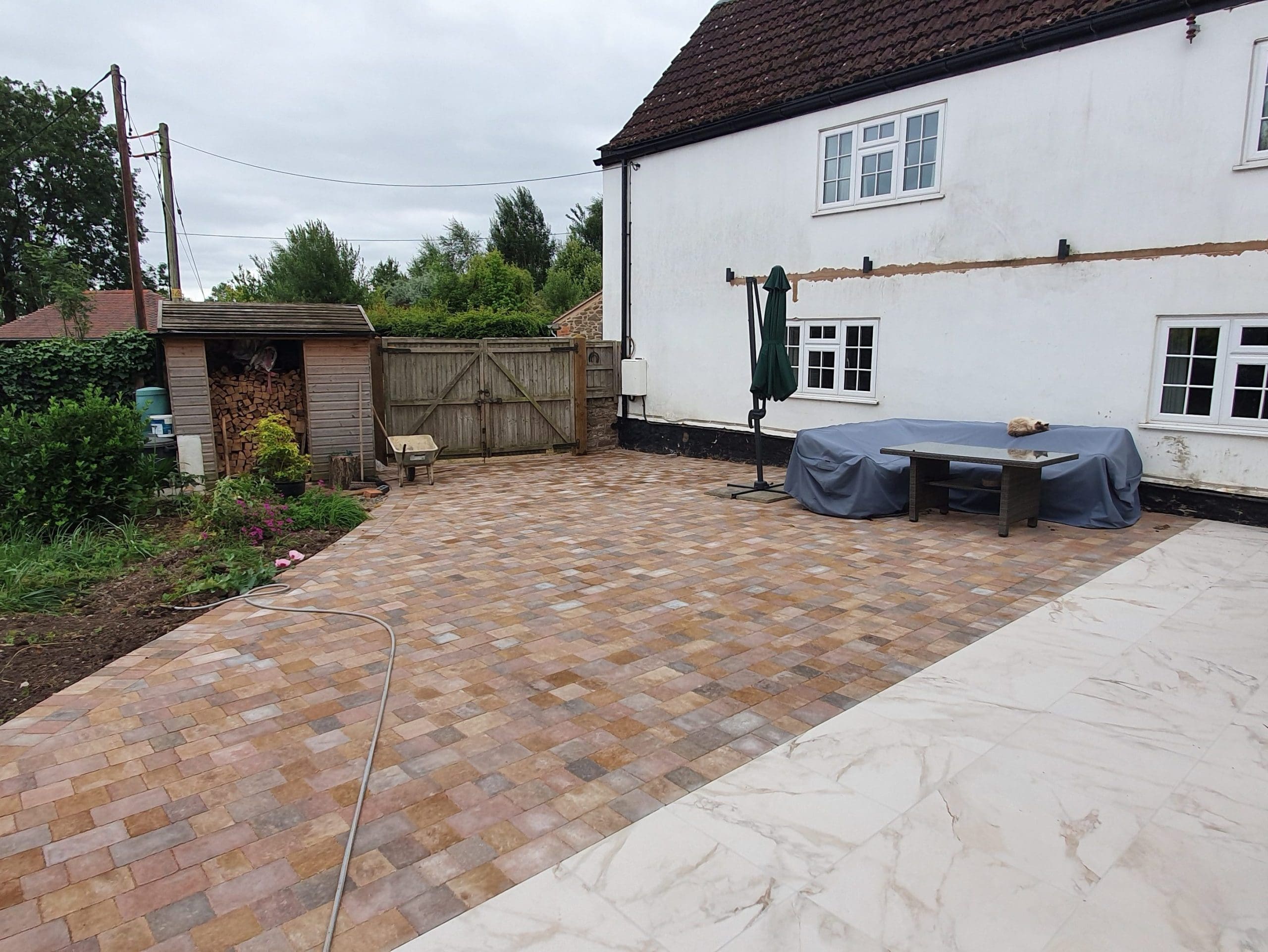 Spacious backyard with a tiled patio, covered table and umbrella near a white house. There’s a grassy garden area, wooden shed, gate, and some potted plants. The sky is overcast.