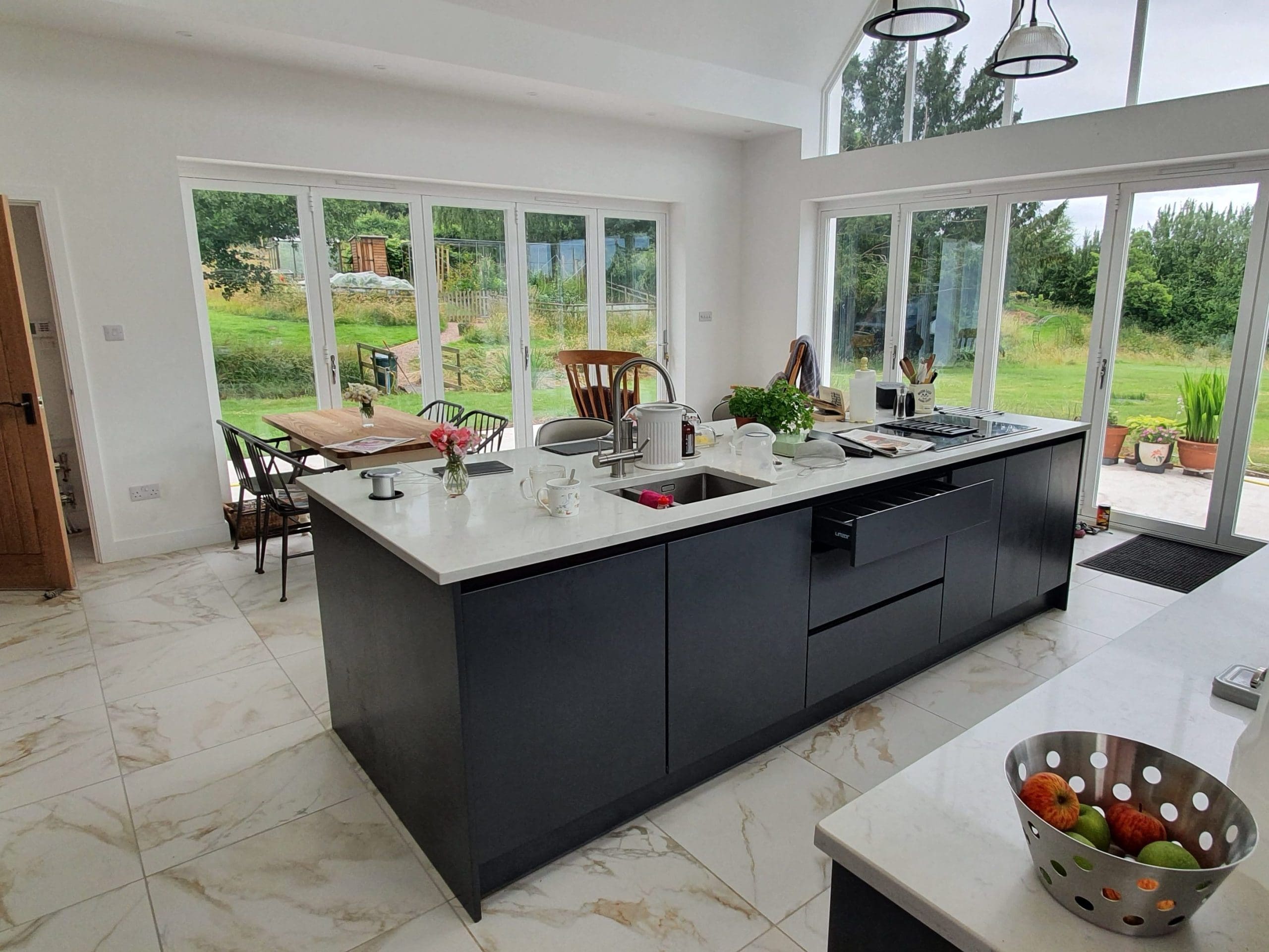 Modern kitchen with large windows, an island with a sink, dark cabinets, white countertops, and a bowl of fruit. To the left is a wooden dining table, and outside, a lush green garden is visible.