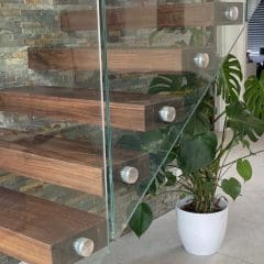 Modern floating wooden staircase with glass railing, next to a stone accent wall. There are potted green plants, including a monstera, on a light tiled floor in a bright, contemporary interior.