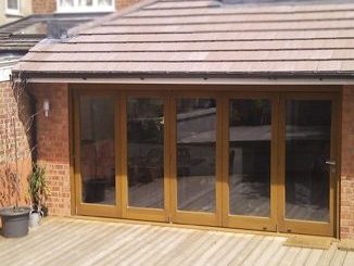 A modern wooden patio door with four glass panels opens to a spacious wooden deck attached to a brick house. Several potted plants are arranged on the left side, and sunlight casts shadows on the deck, creating a warm and inviting atmosphere.