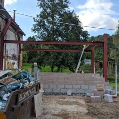 A construction site next to a white house, with steel beams and a partially built concrete block wall. A yellow skip filled with debris is in the foreground, and a red gazebo stands in a grassy area behind.