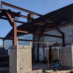 A partially constructed extension with steel beams and concrete block walls is attached to an old house. Two people stand nearby, and building materials are scattered around the site.
