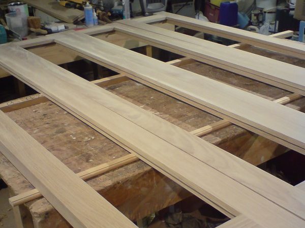 A large wooden frame lies flat on a workbench in a workshop. Various woodworking tools and materials are scattered around the bench. The frame appears to be in the process of assembly, with unfinished wood showing its natural grain patterns.