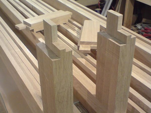 Several wooden planks with precise dovetail joints cut into their ends are arranged on a workbench in a woodworking shop. The background reveals various tools and materials, indicating an active workspace.