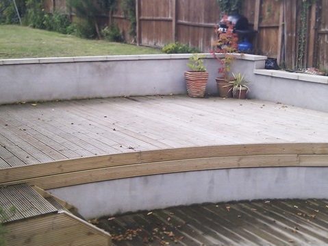 A backyard with a curved wooden deck is shown. The deck has steps leading down to a patio area. There are potted plants placed against the retaining wall on the deck. The yard is surrounded by a wooden fence, with greenery and trees visible in the background.