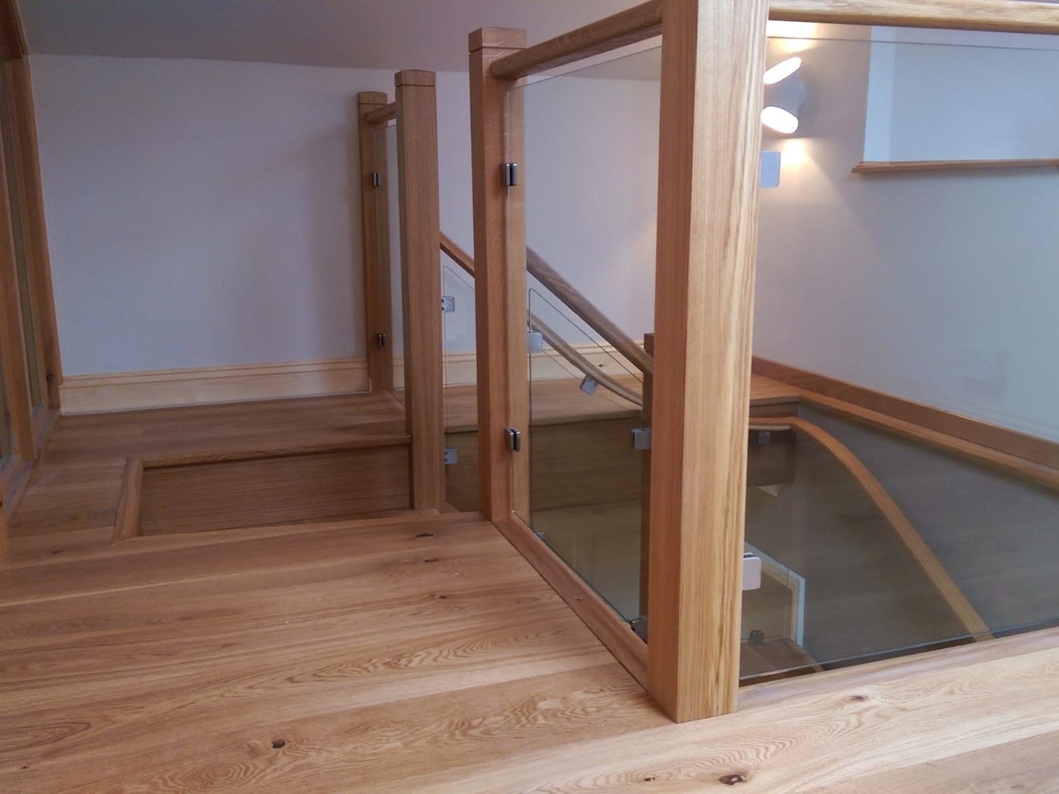 A modern wooden loft floor with glass-paneled railings, leading to a staircase going down. The space is well-lit with light wood tones and minimalist decor.