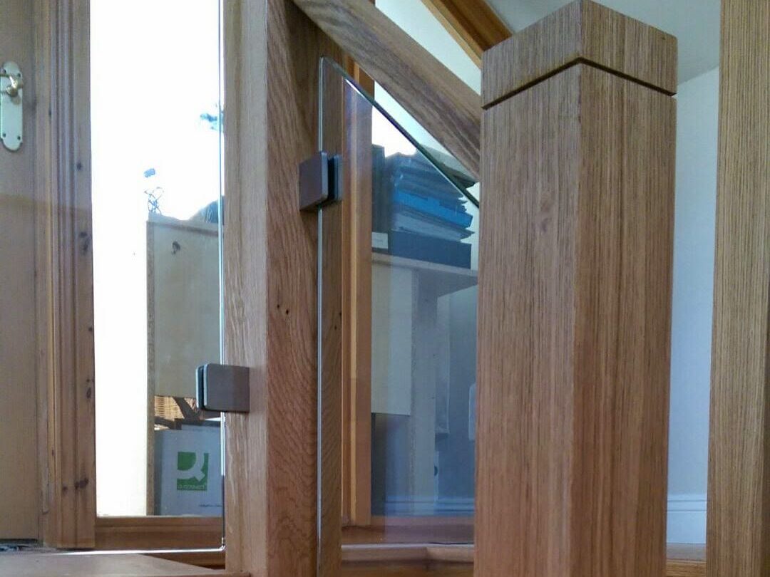A close-up view of a wooden staircase with glass panels in the railing, featuring clean, modern design and natural wood tones. Sunlight streams through large windows, adding warmth to the bright space.